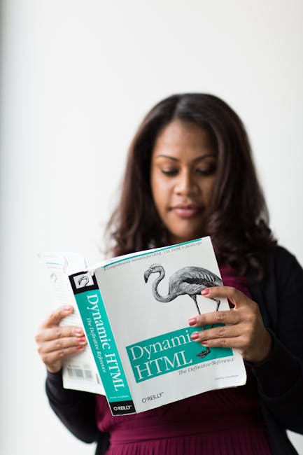 A woman with long, dark hair holding a book titled 'Dynamic HTML' with a flamingo illustration on the cover. She is dressed in a black blazer over a maroon top and appears to be reading or studying th