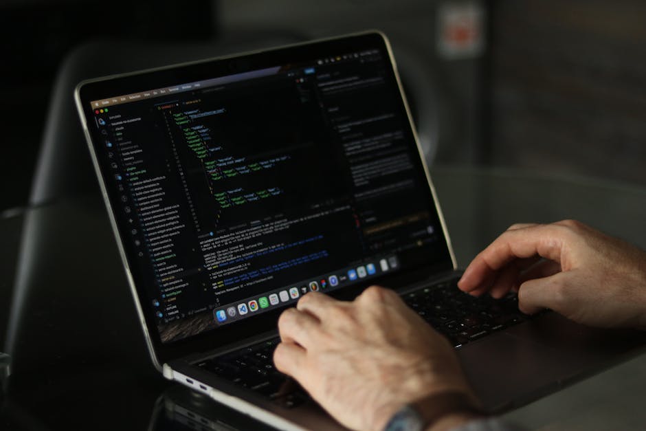 A person working on a laptop with a dark screen displaying lines of code, sitting at a modern table. The laptop has a black keyboard and is slightly open, revealing a vibrant desktop wallpaper and app